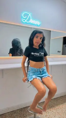 Young woman in a black graphic crop top and denim shorts sitting in a modern salon with neon lighting.