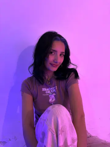 Young woman in a graphic t-shirt sitting against a pink neon lit wall with a moody aesthetic.