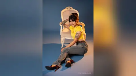 Young woman in a yellow t-shirt and blue jeans sitting on a white vintage chair against a blue studio backdrop.
