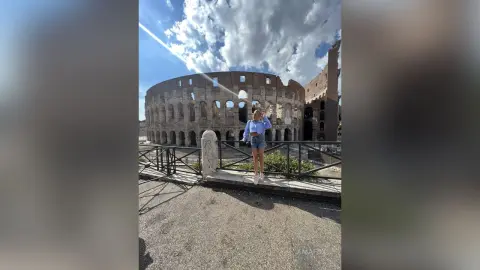 AmaFox — Young woman in a blue shirt and denim shorts standing before the historic Roman Colosseum under a sunny sky.