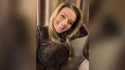 DelliaSmith smiles while wearing a dark snakeskin print top, posing with her hand behind her head against a neutral background.