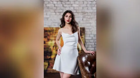Elegant woman in a white satin slip dress posing in a rustic studio with a brick wall background.