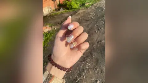 Close-up of a hand with glittery nude manicure and dark beaded bracelet against a natural outdoor background.