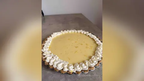 MirandaValentin presents a large, round lemon tart topped with white whipped cream swirls around the edge, sitting on a grey countertop.