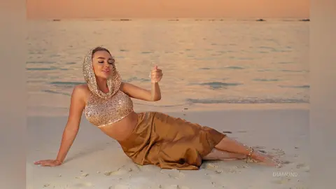 Glamorous woman in a gold sequin hooded crop top and satin skirt relaxing on a tropical beach at sunset.