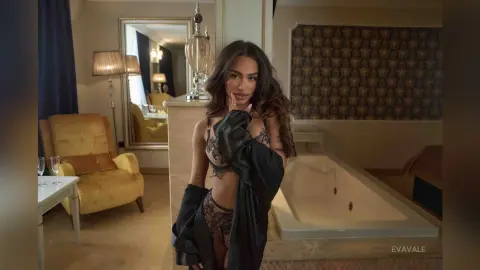 EvaVale poses in black lace lingerie and an open black robe while standing in a luxury hotel room next to a large bathtub.
