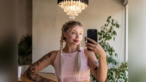 LurlineLacy takes a mirror selfie wearing a pink ribbed tank top, showing off her blonde braids and arm tattoos.