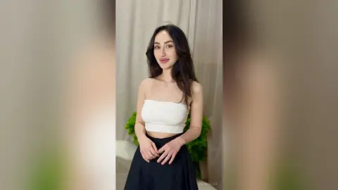 Young woman in a white tube top and black skirt posing elegantly against a neutral indoor background.