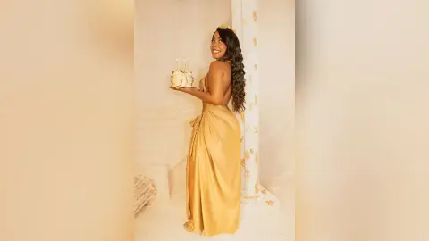 Smiling woman in a gold satin gown holding a birthday cake in a dreamy, elegant white studio setting.