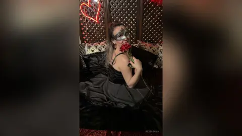 Woman in a black evening gown and masquerade mask holding a red rose in a romantic, moody setting.