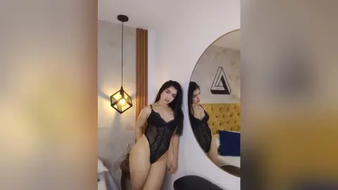 Young woman in black lace lingerie posing confidently in a modern minimalist bedroom setting.