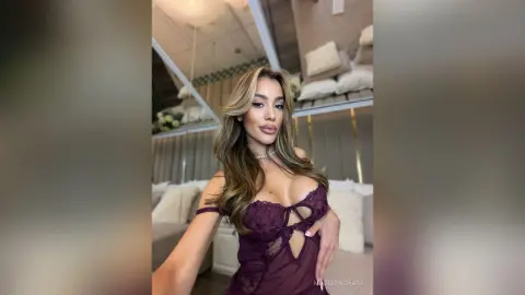 Glamorous woman in a burgundy lace bodysuit posing in a luxury modern bedroom setting.