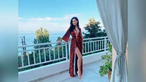Woman in orange tiger print robe standing on a sunny balcony overlooking a lush coastal landscape.