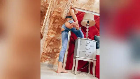 ChristineDavis poses naked from the waist down, wearing blue denim jeans that expose her ass while she bends over next to a red headboard and lamp.