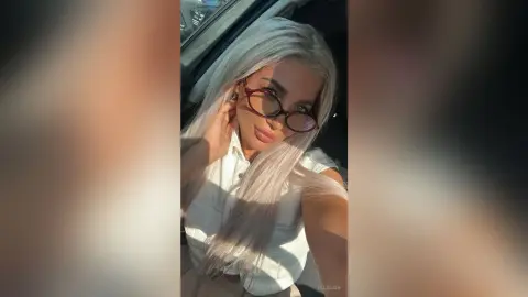Blonde woman in a white button-down shirt wearing glasses, posing with a soft glow inside a car.