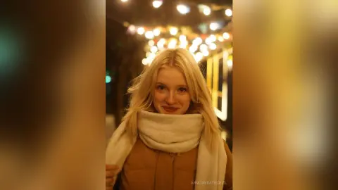 Blonde woman in a cozy white scarf and brown coat smiling under warm festive lights at night.