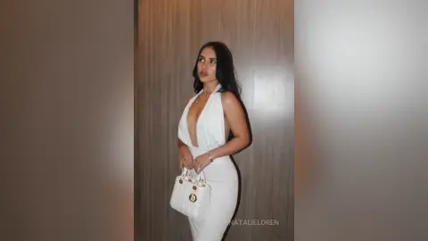 Elegant woman in a white plunging halter dress holding a designer handbag against a neutral wood background.