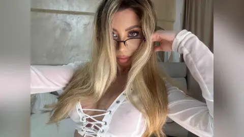 Blonde woman in a white sheer lace-up top posing in a bright bedroom setting.