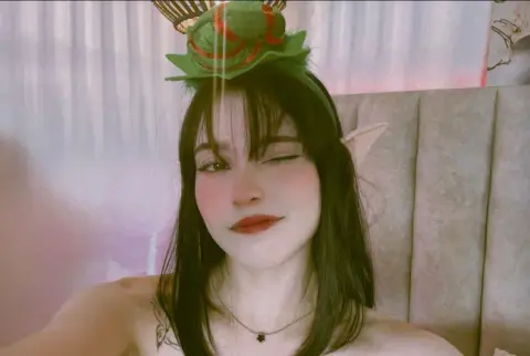 Young woman with dark hair and elf ears wearing a green leafy headpiece in a soft, dreamy indoor setting.