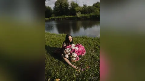 Young woman in a dark dress holding a large pink bouquet by a peaceful lake in a lush green meadow.