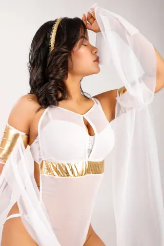 Woman in white sheer bodysuit and gold accents posing gracefully in ethereal flowing fabric.
