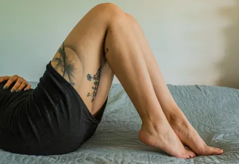 Woman in black skirt relaxing on a bed with visible leg tattoos in a peaceful indoor setting.