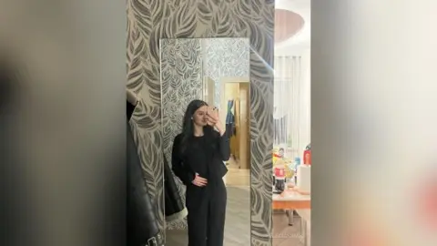 Young woman in a chic black outfit taking a mirror selfie in a modern room with patterned wallpaper.