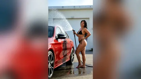 Smiling woman in a black bikini washing a red car with a high-pressure water hose outdoors on a sunny day.