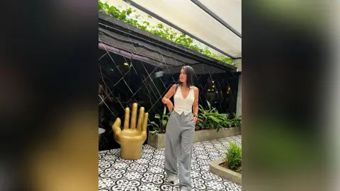 Stylish woman in a white sleeveless top and grey trousers standing in a modern indoor garden setting.