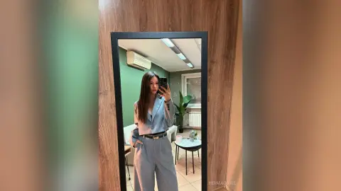 Young woman in a light blue oversized suit taking a mirror selfie in a modern minimalist room.