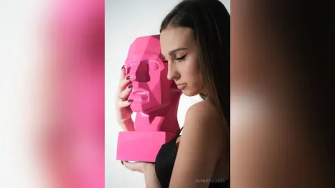 Young woman in a black top posing thoughtfully with a pink geometric sculpture in a minimalist studio setting.