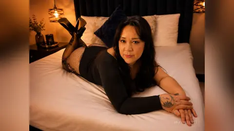 Curvy woman in black bodysuit and fishnet tights relaxing on a white bed in a cozy, dimly lit bedroom.