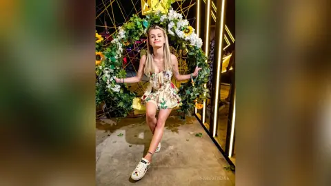 Blonde woman in a floral summer dress posing joyfully in front of a large white flower wreath at night.