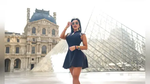Dolce stands outdoors in a black sleeveless mini dress and sunglasses, posing in front of the glass Louvre pyramid.