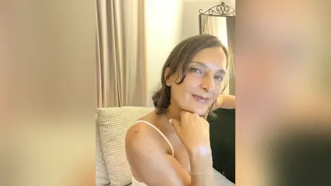 MichaelaBelle smiles while resting her chin on her hand, wearing a white camisole or tank top top.