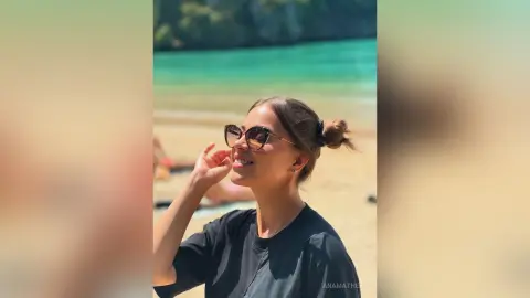 AnaMathers smiles while wearing sunglasses and a black t-shirt, with her hair tied in a bun at a sunny beach.