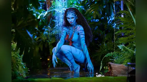 Blue Na'vi woman with long dark hair crouching in a lush tropical jungle near a misty waterfall.