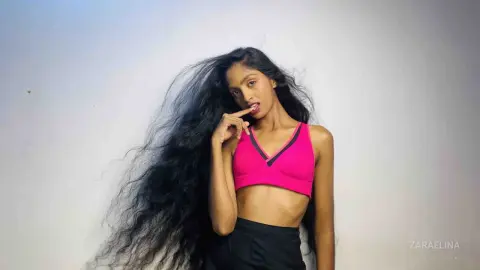 Young woman with long wavy hair wearing a pink sports bra and black skirt in a studio setting.