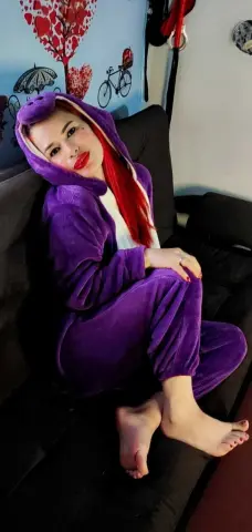 Red-haired woman in a cozy purple fleece onesie relaxing on a black sofa in a bright room.
