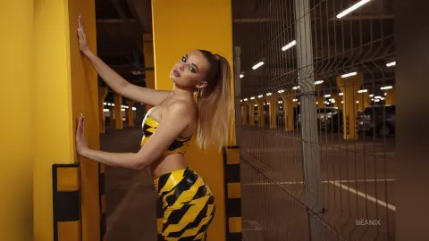 BeaNix poses against a bright yellow pillar wearing a yellow and black striped bikini set and long blonde hair.