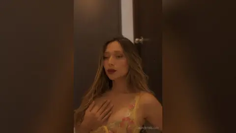 LaurentBelluci poses with her hand resting on her chest, wearing a yellow floral lace bra or camisole.