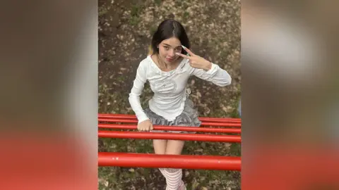 MielVerane wearing a white long-sleeve shirt and a grey skirt while posing behind red metal bars outdoors.