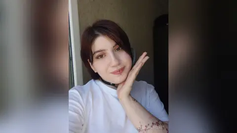 OrianaSecret — Smiling young woman with short brown hair wearing a white t-shirt in a bright indoor setting.