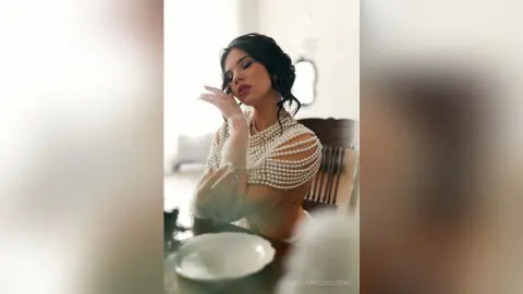 JenellDelorme poses with her hands near her face, wearing a white pearl-beaded top that shows her cleavage and shoulders, with her dark hair styled back.