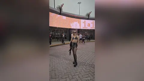 GiovannaKlein stands outdoors in a black crop top and sheer leggings, posing in front of a large building with a digital display.