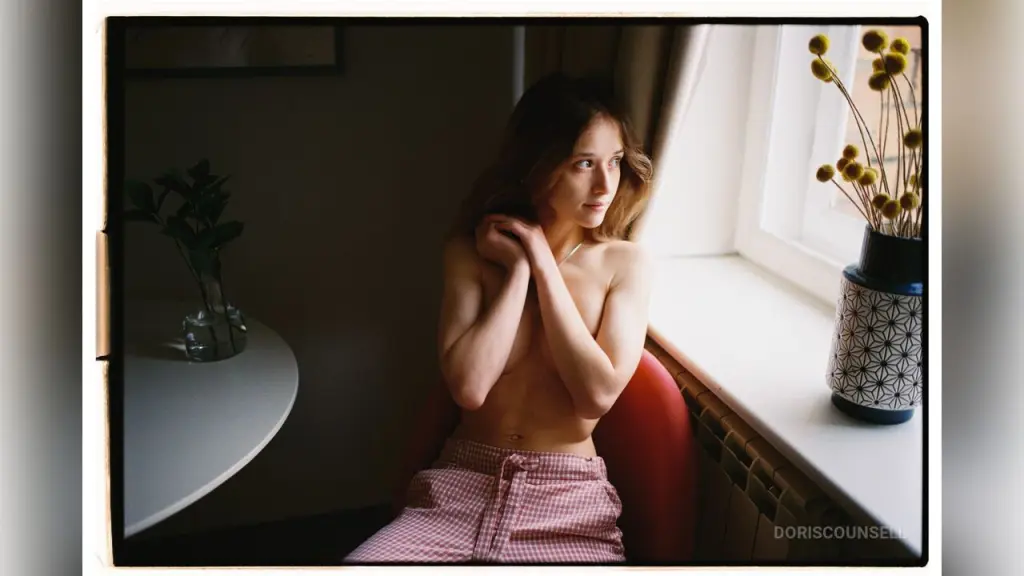 Smiling blonde woman in a white eyelet blouse posing in a soft, moody indoor setting.