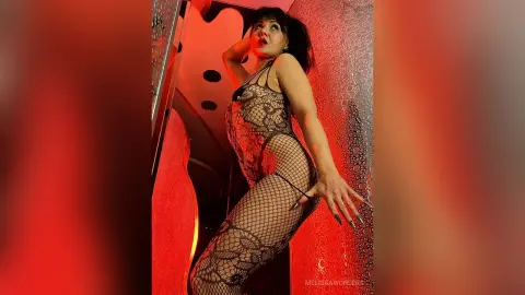 Glamorous woman in a black lace bodysuit posing in a moody, red-lit modern interior.