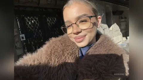 Smiling young woman in a cozy brown faux fur coat and glasses outdoors in bright sunlight.
