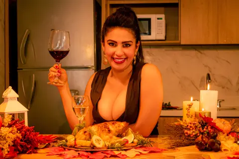 Smiling woman in a black dress holding red wine at a festive Thanksgiving dinner with roasted turkey and candles.