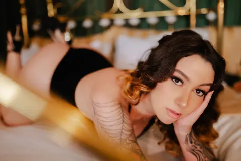 spicyspiders21 poses on a bed wearing a black bodysuit, showing off her dark wavy hair and large arm tattoos while looking at the camera.